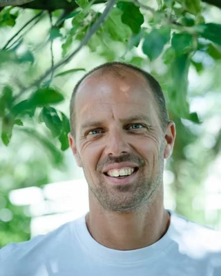 Een foto van Jeroen in een lichtblauw T-shirt, tegen een groene achtergrond, waarin enkele bladeren zichtbaar zijn.