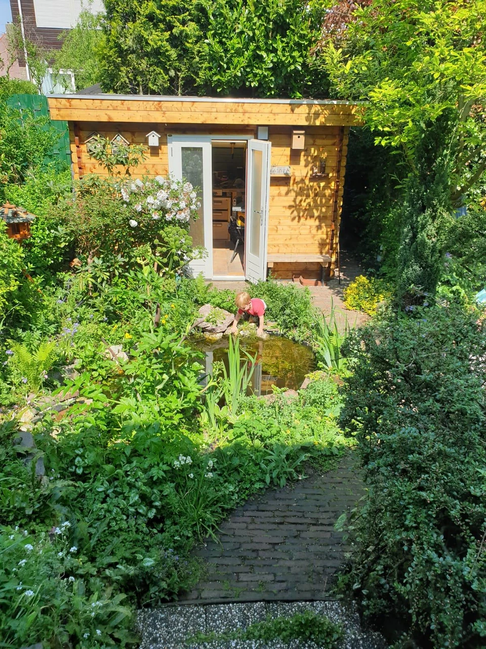 Een foto van een paadje dat richting een schuur loopt, met in het midden van de tuin een vijfer met daarnaast een spelend kind.