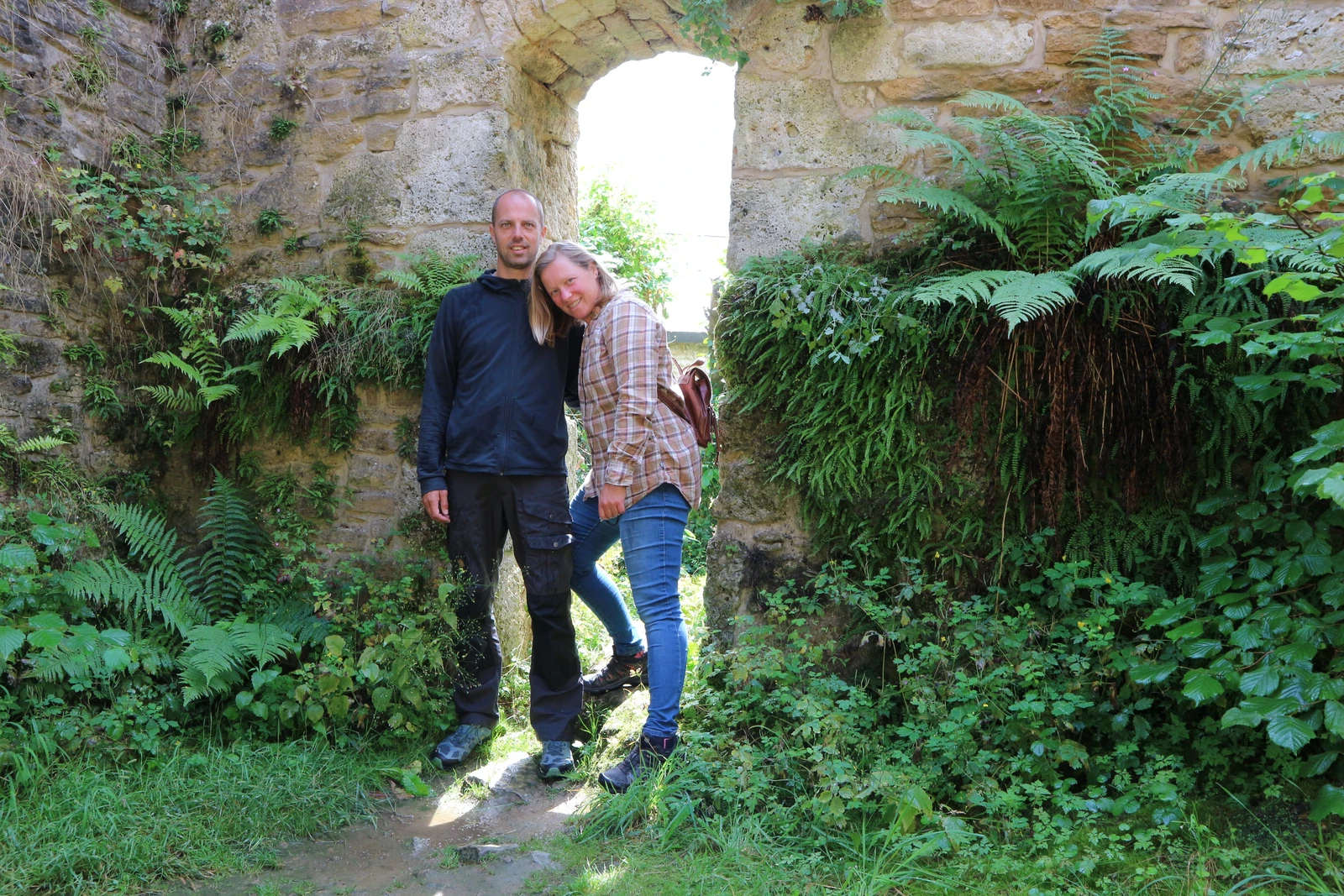 Een foto van Jeroen en Mirjam voor een poort in een grote lichtbruine stenen muur. Ze dragen wandelkleding en tegen de muur groeien verschillende grote planten.