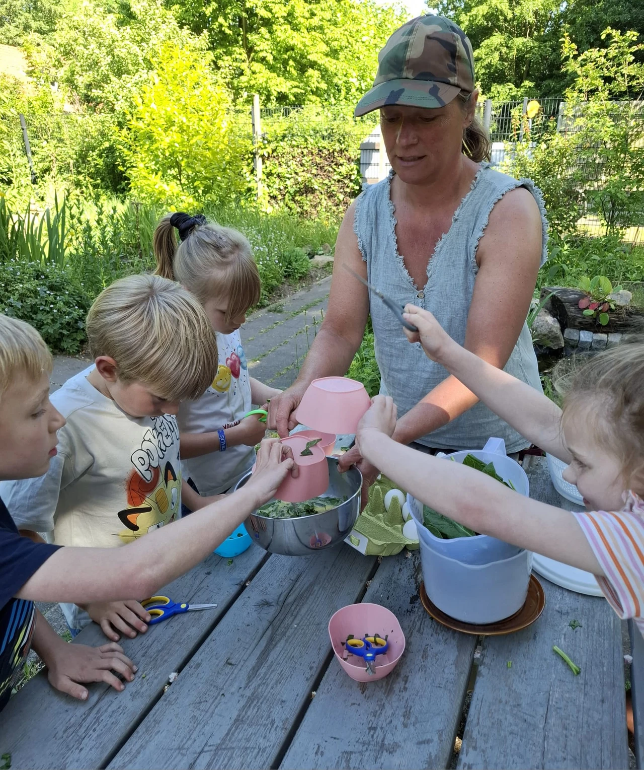Een foto van Mirjam die met een groepjes kinderen in een tuin een salade maakt. Ze houdt een metalen kom vast waar de kinderen roze bakjes met ingrediënten in legen. Op tafel staat een doos eieren en nog meer bakjes. Op de achtergrond zie je verschillende bedden met kruiden.